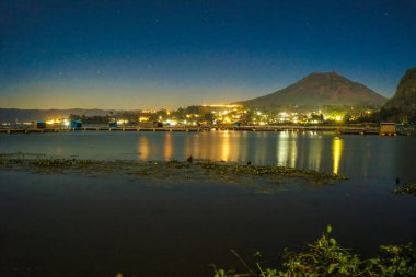 Batur Gölü, Kintamani, Bali 'deki akşam atmosferi, Batur Dağı' nın yansıması ve göl kenarındaki köyün atmosferi parlak ışıklarla aydınlatılıyor. Gece Fotoğrafçılığı.