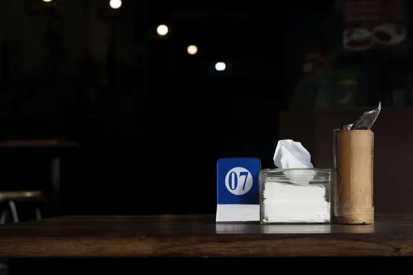 A wooden table with table number, a box of tissues and a bamboo cutlery holder, illuminated by a single light source in the background.