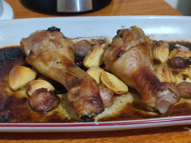 İki kızarmış tavuk budu, kahverengi derili ve çıtır çıtır kenarlı bir tabakta. Sarımsaklar arka planda görünüyor..