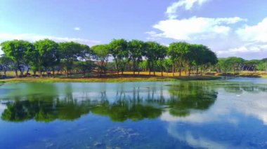 Bir dron durgun bir gölün üzerinde süzülür, kamerası parıldayan yüzey boyunca döner. Kıyıdaki ağaçlar rüzgarda sallanıyor yaprakları masmavi gökyüzüne karşı parlak bir yeşil. İnsansız hava aracı görüntüleri
