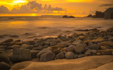 Papuma, Jember, Doğu Java, Endonezya 'daki kayalık sahilde gün batımı. Okyanus dalgalarıyla dolu bulutlu bir öğleden sonra kumları aşındırıyor. Doğa ve Manzara Fotoğrafçılığı.