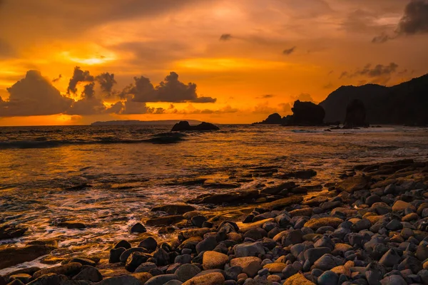 Papuma, Jember, Doğu Java, Endonezya 'daki kayalık sahilde gün batımı. Okyanus dalgalarıyla dolu bulutlu bir öğleden sonra kumları aşındırıyor. Doğa ve Manzara Fotoğrafçılığı.