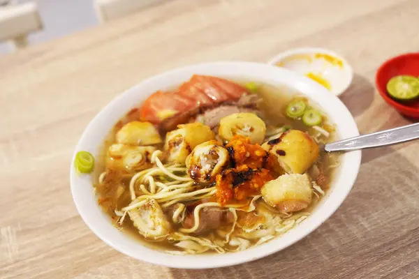 A steaming bowl of Soto Mie Bogor, a traditional Indonesian noodle soup, featuring thin yellow noodles, tender chunks of beef, crisp wonton skins, and fresh vegetables