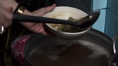 Bir insanın elleri, metal bir tencereden kepçeyle beyaz bir seramik kaseye et ve sakatat çorbası dökerken görülüyor. Surabaya, Endonezya 'da.