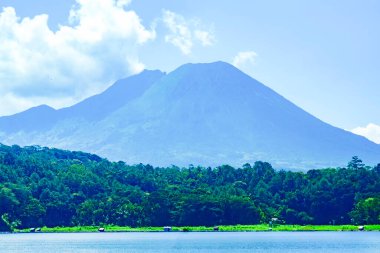 a majestic of Mount Lemongan with a calm lake in front of it. Lush trees surround the base of the volcano, creating a peaceful scene in Lumajang, East Java, Indonesia. clipart