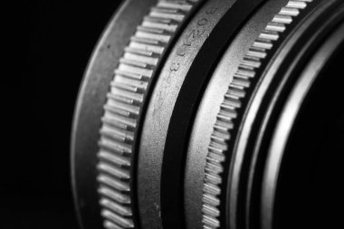 Görüntü, bir kamera lensinin siyah-beyaz yakın çekimi. Objektifin birkaç kavisli cam elementi ve metal bir yuvası var..