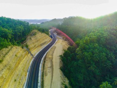 JLS ya da South Coast Road, Tulungagung, Doğu Java, Endonezya 'daki sık yeşil bir ormanın içinden geçen kıvrımlı bir yol. Güneş ışığı ağaçların yapraklarının arasından süzülüyor, yol üzerindeki noktalı ışığı yansıtıyor.