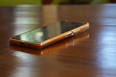 Parlak altın renkli akıllı telefon ahşap bir masanın üzerinde duruyor, ışığı yansıtıyor ve ince bir parıltı yayıyor..