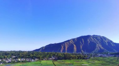 Sembalun Tepeleri, Lombok, Endonezya 'nın nefes kesici hava manzarası. Berrak mavi gökyüzü altında yemyeşil alanlar, sakin bir köy ve görkemli dağlar..