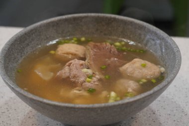 Bir kase baharatlı bakso çorbası köfte, erişte ve et suyu, yanında küçük bir tabak limon dilimi ve biber sosu, yanında da bir kase vermicelli şehriye ve küçük bir tabak kızarmış arpacık soğanı..