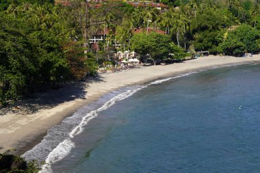 Pantai Senggigi, Lombok, Endonezya yakınlarındaki Idyllic plaj sahnesinde el değmemiş beyaz kum, yumuşak dalgalar, yemyeşil ve palmiye ağaçlarıyla dolu bir sahil beldesi yer alıyor..