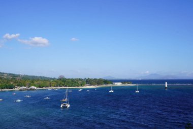 Pantai Senggigi, Lombok, Endonezya 'nın nefes kesen panoramik manzarası, el değmemiş bir sahil, turkuaz sular, demirlemiş yelkenliler ve yemyeşil tepeler ve açık mavi gökyüzü karşısında beyaz bir deniz feneri sergileniyor..
