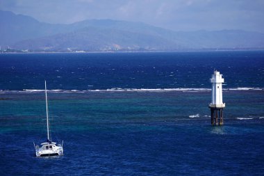 Pantai Senggigi, Lombok, Endonezya yakınlarındaki deniz manzarasında kayalık bir çıkıntının üzerinde duran beyaz bir deniz feneri, turkuaz sularda demirlemiş bir yelkenli ve berrak mavi gökyüzünün altındaki uzak dağlar yer alıyor..