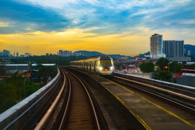 Yükseltilmiş bir tren, günbatımında hareketli bir şehirden geçer, Kuala Lumpur, Malezya yakınlarındaki arka planda gökdelenler ve otoyollar vardır..