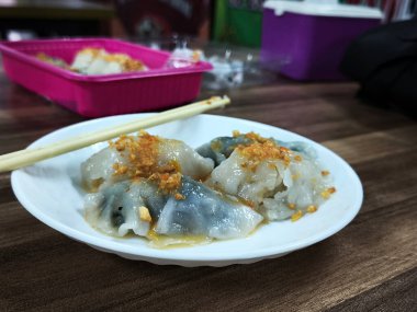 A plate of Choipan cakes from Indonesia sits on a wooden table. The cakes are white with green accents and are topped with a red sauce. Chopsticks are placed on the plate. clipart