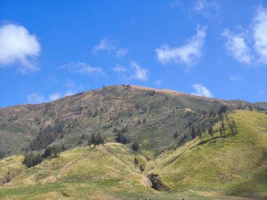 Bromo Tengger Semeru Ulusal Parkı 'nın görkemli manzarası. İkonik Bromo Dağı kendine özgü krateriyle dimdik ayakta duruyor. Etrafı geniş bir volkanik kum ve açık mavi bir gökyüzü ile çevrili.