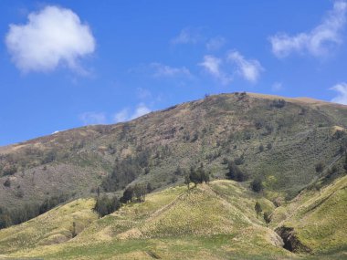 Bromo Tengger Semeru Ulusal Parkı 'nın görkemli manzarası. İkonik Bromo Dağı kendine özgü krateriyle dimdik ayakta duruyor. Etrafı geniş bir volkanik kum ve açık mavi bir gökyüzü ile çevrili.