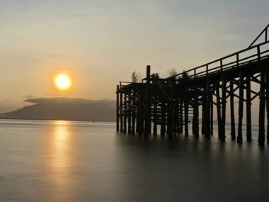 Gündoğumunda, Endonezya 'nın Banyuwangi kenti yakınlarındaki destek direklerine yapışmış, yıpranmış ahşap bir iskele suya doğru uzanıyor.