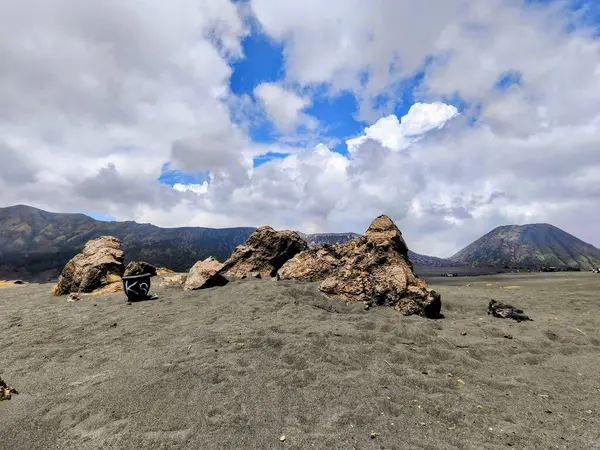Endonezya 'daki Bromo Dağı' nın nefes kesici manzarası. Volkanın zirvesi dramatik bulutlarla örtülmüş, korku ve gizem duygusu yaratıyor. Ön planda kayalık bir manzara var.