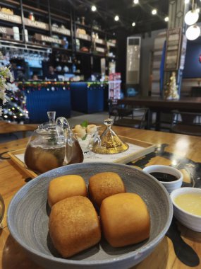 Ahşap bir masada taze pişmiş çöreklerle dolu rahat bir kafe sahnesi. Ayrıca bir çaydanlık ve bir bardak çay, bir vazo dolusu çiçek ve bir tepsi dolusu meyve var..