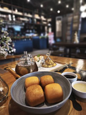 Ahşap bir masada taze pişmiş çöreklerle dolu rahat bir kafe sahnesi. Ayrıca bir çaydanlık ve bir bardak çay, bir vazo dolusu çiçek ve bir tepsi dolusu meyve var..