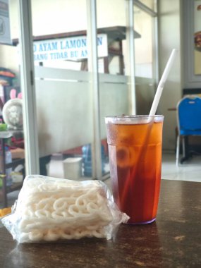 A refreshing iced tea with a straw and a bag of Kerupuk, Indonesian crackers on a table, ready to be enjoyed as a snack. clipart