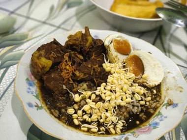 A bowl of Rawon, a rich and flavorful Indonesian beef stew with a dark broth, served with bean sprouts, a boiled egg, and crackers. clipart