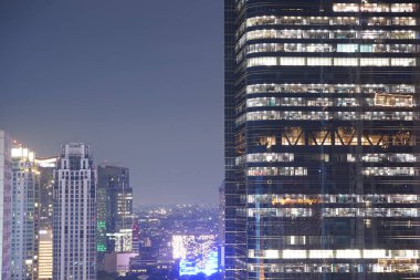 Kalabalık şehir siluetinin gece manzarası. Yüksek bir gökdelen çerçevenin sağ tarafına hükmediyor. Aydınlatılmış pencereleri karanlık gece gökyüzü ile çelişen ızgara benzeri bir desen oluşturuyor..