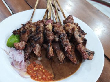 Bir tabak Sate Kambing (Keçi Satay). Şişler, beyaz bir tabakta düzgünce yerleştirilmiş ızgara keçi eti dolu. Sataya bir kase fıstık sosu eşlik eder, küçük bir kase sambal. 