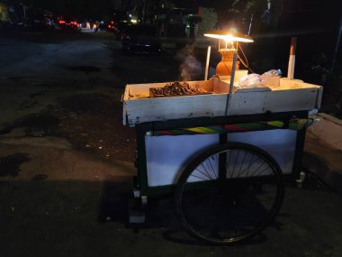 Çekici seyyar satıcı sıcak bir lambanın altında fıstık servis ediyor. Gece aşermeleri için mükemmel. Lezzetli lokmalar için rahat bir yer.