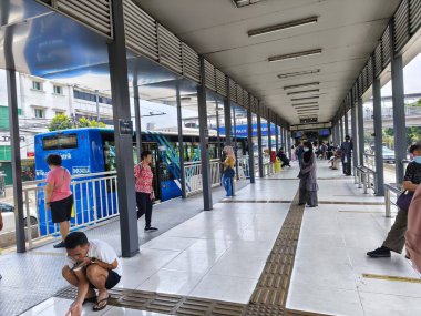 1 Mart 2025. Jakarta, Endonezya. Trans Jakarta otobüs durağında hareketli bir sahne, üç kişinin yürüdüğü, canlı bir atmosferi ve arka planda açık tabelaları olan.