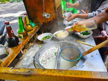 Bir sokak yemeği satıcısı mie ayam veya Endonezya usulü köfteli erişte çorbası, taze yeşillik ve et suyu hazırlar, yağlı ve kepçeli sıcak çorbayı kırsal bir tezgahta kaselere koyar..
