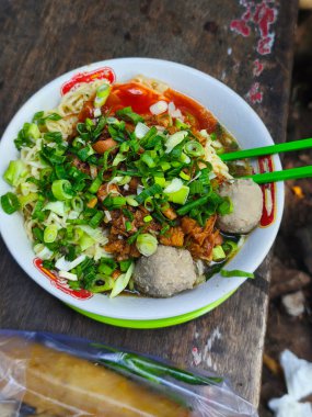 Bir kase lezzetli Mie Ayam ya da doğranmış yeşil soğan, köfte ve baharatlı sos ile kaplı Endonezya usulü tavuk çorbası, kırsal ahşap bir masada yeşil çubuklarla servis edilir..