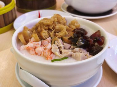 Kızarmış hamur çubukları, karides, tavuk ve korunmuş siyah yumurtalarla kaplı bir kase pirinç lapası. Tahta bir masada kaşıkla servis edilen beyaz bir kasede..