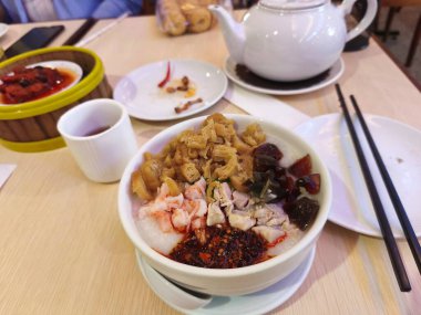 Kızarmış hamur çubukları, karides, tavuk ve korunmuş siyah yumurtalarla kaplı bir kase pirinç lapası. Tahta bir masada kaşıkla servis edilen beyaz bir kasede..