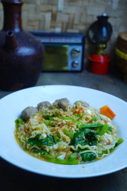 Bir kase Endonezya usulü hazır erişte köfte, sebze ve kızarmış arpacık soğanıyla servis edilir, arka planda antika dekoru olan ahşap bir masaya yerleştirilir..