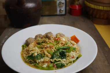 Bir kase Endonezya usulü hazır erişte köfte, sebze ve kızarmış arpacık soğanıyla servis edilir, arka planda antika dekoru olan ahşap bir masaya yerleştirilir..