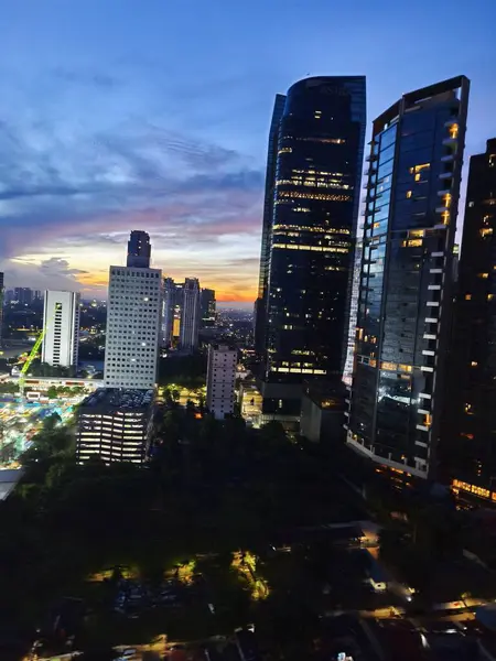 Jakarta 'nın canlı şehir manzarası alacakaranlıkta aydınlık gökdelenler, parlayan bir günbatımı gökyüzü ve aşağıdaki hareketli şehir caddeleri.