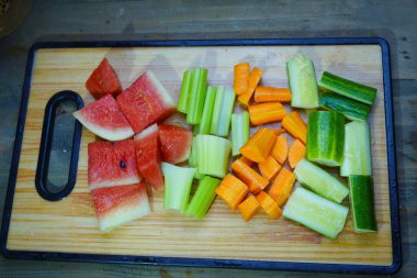 Çeşitli kesilmiş sebzeler ve karpuz parçaları ahşap bir kesme tahtasına dizilmiştir.