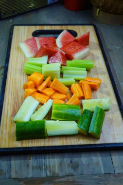 Çeşitli kesilmiş sebzeler ve karpuz parçaları ahşap bir kesme tahtasına dizilmiştir.