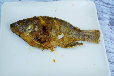 Mükemmel kızartılmış bir Mujair ya da Tilapia beyaz dikdörtgen bir tabakta duruyor ve arka planda küçük bir kase koyu sos var..