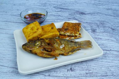 Bir tabak altın kaplama Mujair veya Tilapia, tofu ve tempeh içerir, arka planda küçük bir kase koyu sos, geleneksel Endonezya yemeği sergilenir