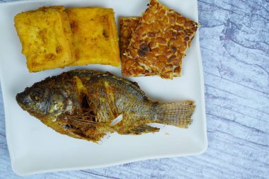Bir tabak altın kaplama Mujair veya Tilapia, tofu ve tempeh içerir, arka planda küçük bir kase koyu sos, geleneksel Endonezya yemeği sergilenir