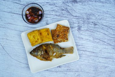 Bir tabak altın kaplama Mujair veya Tilapia, tofu ve tempeh içerir, arka planda küçük bir kase koyu sos, geleneksel Endonezya yemeği sergilenir