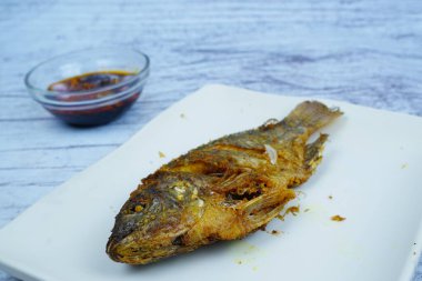 Mükemmel kızartılmış bir Mujair ya da Tilapia beyaz dikdörtgen bir tabakta duruyor ve arka planda küçük bir kase koyu sos var..