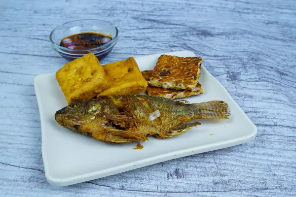Bir tabak altın kaplama Mujair veya Tilapia, tofu ve tempeh içerir, arka planda küçük bir kase koyu sos, geleneksel Endonezya yemeği sergilenir