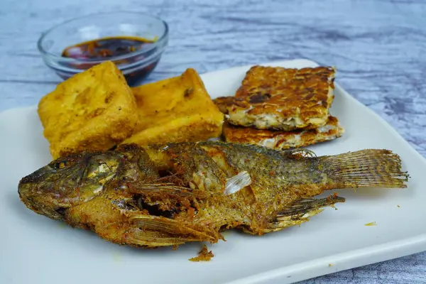 Bir tabak altın kaplama Mujair veya Tilapia, tofu ve tempeh içerir, arka planda küçük bir kase koyu sos, geleneksel Endonezya yemeği sergilenir