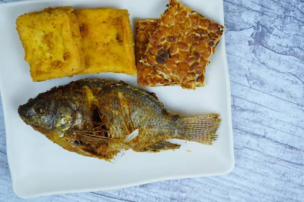 Bir tabak altın kaplama Mujair veya Tilapia, tofu ve tempeh içerir, arka planda küçük bir kase koyu sos, geleneksel Endonezya yemeği sergilenir