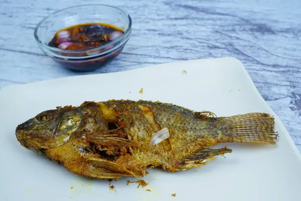 Mükemmel kızartılmış bir Mujair ya da Tilapia beyaz dikdörtgen bir tabakta duruyor ve arka planda küçük bir kase koyu sos var..