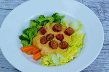 Beyaz tabakta pişmiş brokoli, lahana, havuç ve kremalı sarı sosun üzerinde dilimlenmiş sosisler var..